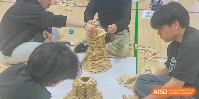 Students gather around a white sheet on the floor to build a tall, tapering tower out of wooden blocks, demonstrating teamwork and precision at the Korean International School In Beijing.