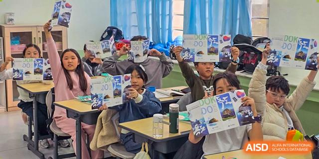 A classroom full of students smiles while holding up their completed educational brochures to the camera at the end of a successful lesson at the Korean International School In Beijing.