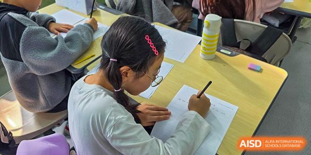 A young student focuses intently while writing on a worksheet at a wooden desk in a bright classroom setting during a lesson at the Korean International School In Beijing.