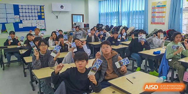 A group of students sit at their desks, proudly holding up blue pamphlets and smiling toward the camera during a special activity at the Korean International School In Beijing.