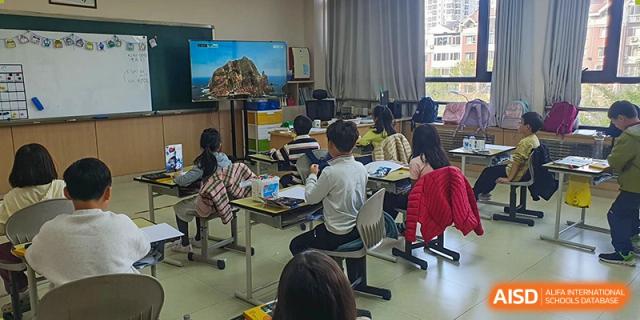 A wide view of a classroom shows students seated at desks facing a large screen displaying a coastal landscape during a lecture at the Korean International School In Beijing.