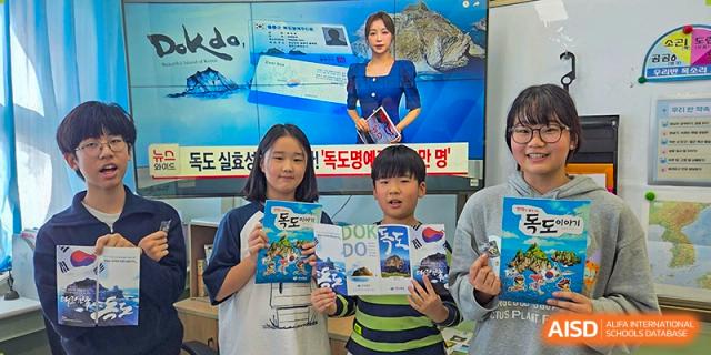 Three cheerful students stand together in front of a digital screen, holding up educational booklets about Dokdo to the camera at the Korean International School In Beijing.