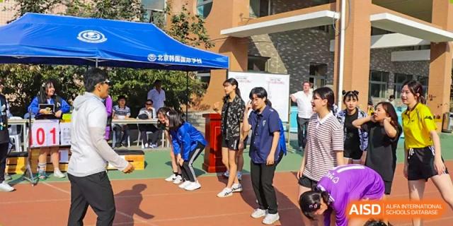 Students gather outdoors on a sports court near a blue tent, participating in a physical education activity or sports day event at the Korean International School In Beijing.