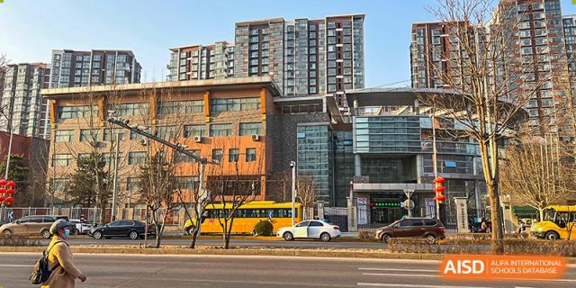 A yellow school bus is parked on a city street in front of the multi-story grey and brown stone academic buildings belonging to the Korean International School In Beijing.