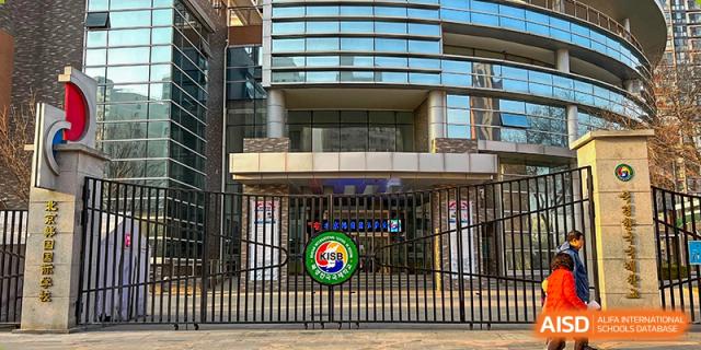 A close-up view shows the modern glass entrance and black gated fence featuring a colorful circular school emblem at the main gate of the Korean International School In Beijing.