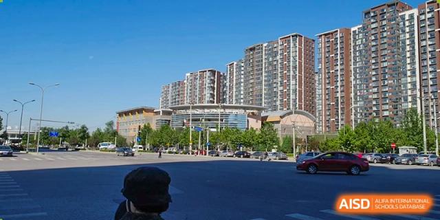 A wide perspective from across the street captures the expansive campus architecture set against a backdrop of tall residential towers at the Korean International School In Beijing.