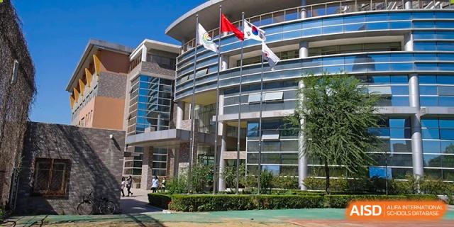 Three flags, including the South Korean national flag, fly prominently on tall silver poles in front of the contemporary glass facade of the Korean International School In Beijing.