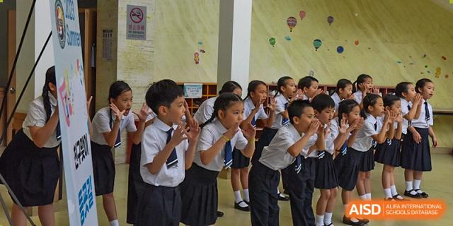 A row of young students in school uniforms lean forward and cup their hands around their mouths as if shouting or cheering together at the Korean International School In Beijing.