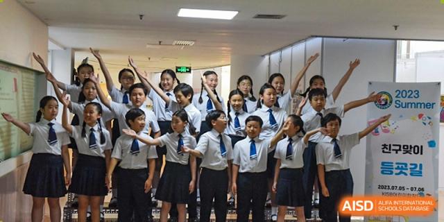 Cheerful students in matching school uniforms raise their arms and sing joyfully during a choir performance in a bright, modern indoor hallway at the Korean International School In Beijing.