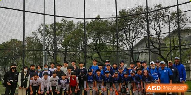 A large group of students wearing baseball uniforms and casual athletic gear pose together for a team photo on an outdoor sports field at the Korean International School In Beijing.
