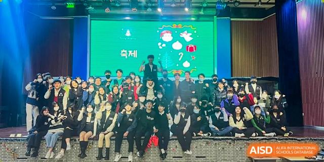 A large group of students and teachers pose together on a brightly lit stage in front of a green digital screen during a festive holiday celebration at the Korean International School In Beijing.