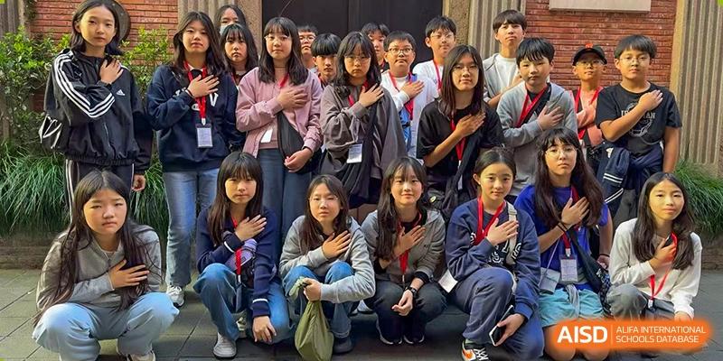 A large group of students posing together outdoors, many with their hands over their hearts, wearing lanyards at Korean International School In Beijing.