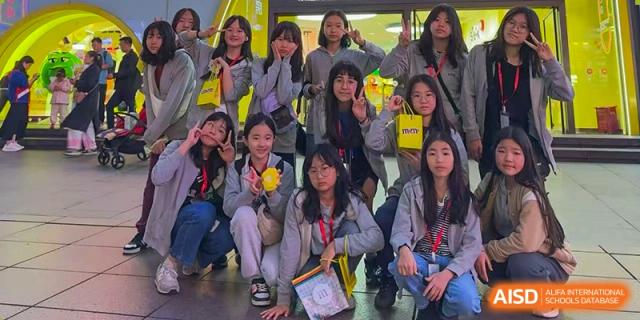 A group of female students smiling and posing for a group photo while holding yellow gift bags indoors at Korean International School In Beijing.