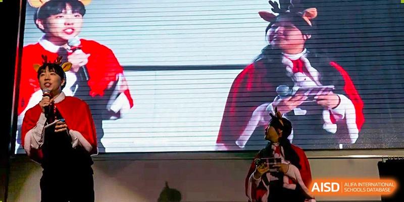 A student dressed in a festive red holiday costume with reindeer antlers stands in front of a large screen at Korean International School In Beijing.