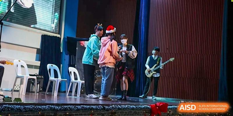 Students performing a skit or musical number on a stage decorated with holiday hats and musical instruments at Korean International School In Beijing.