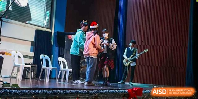 Students performing a skit or musical number on a stage decorated with holiday hats and musical instruments at Korean International School In Beijing.