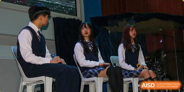 Two female students in school uniforms sitting on stage and participating in a presentation or performance at Korean International School In Beijing.