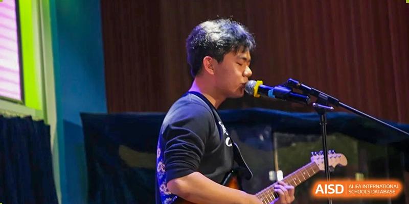 A student singing into a microphone while playing an electric guitar during a school talent show or concert at Korean International School In Beijing.