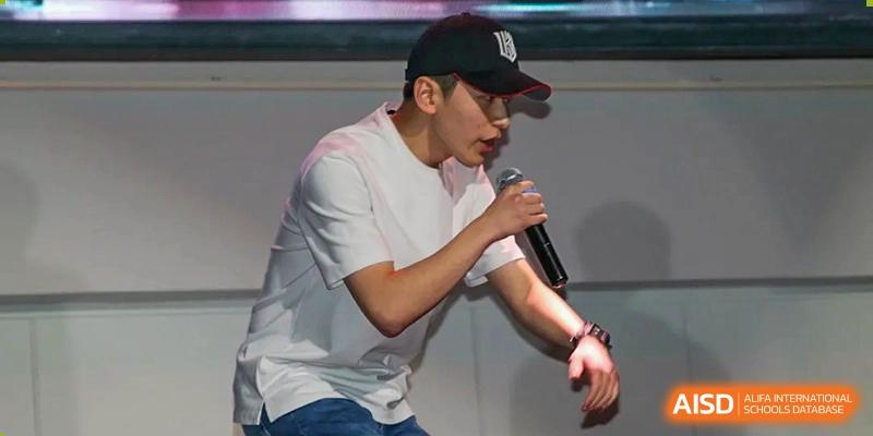 A young man in a white t-shirt and baseball cap performing a vocal piece or rap on stage during an event at Korean International School In Beijing.