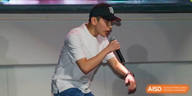 A young man in a white t-shirt and baseball cap performing a vocal piece or rap on stage during an event at Korean International School In Beijing.