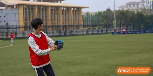 A player in a red vest and white shirt catches a ball on a green turf baseball field during practice at Korean International School In Beijing.