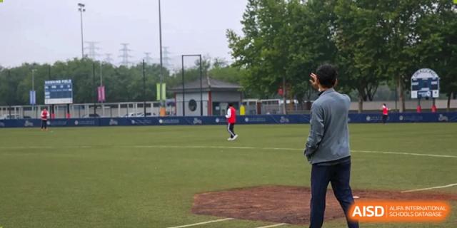 A baseball player in a gray shirt stands on the field looking toward the pitcher's mound during a game at Korean International School In Beijing.