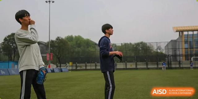 A student in a navy hoodie and black track pants stands ready with a baseball glove on a green field at Korean International School In Beijing.