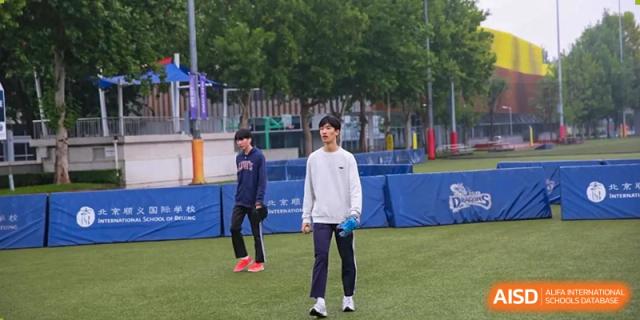 A male student wearing a white sweatshirt and dark athletic pants stands on the green field during an outdoor sports practice session at the Korean International School In Beijing.