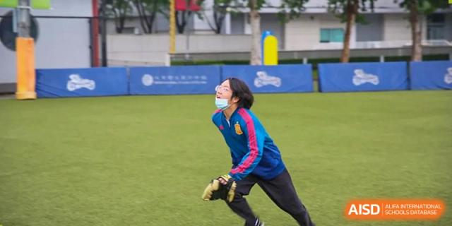 A young man wearing a blue and red tracksuit smiles while looking up and preparing to catch a ball during a bright day of outdoor activities at the Korean International School In Beijing.
