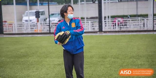 A student wearing a face mask and a blue athletic jacket stands ready with a baseball glove on a green field during physical education at the Korean International School In Beijing.