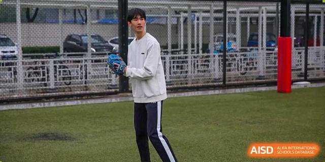 A student in a white long-sleeve shirt and track pants holds a blue baseball glove while focusing on the game during an outdoor sports event at the Korean International School In Beijing.