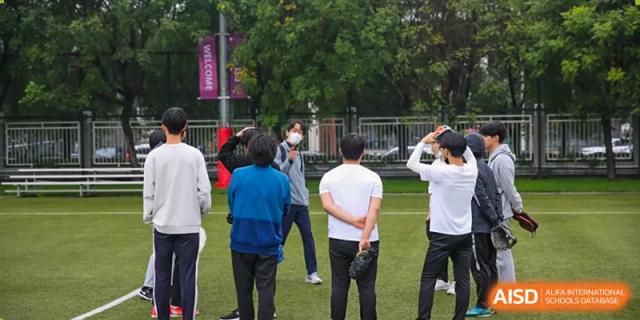 A group of students in casual athletic wear gathers in a circle on the grass to discuss team strategy before a game begins at the Korean International School In Beijing.