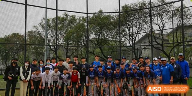 A large group of students in blue and grey baseball uniforms poses together for a team photo in front of a green fence at the Korean International School In Beijing.