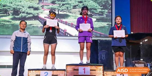 Three students stand proudly on a wooden awards podium holding certificates and wearing medals after a successful competition held at the Korean International School In Beijing.
