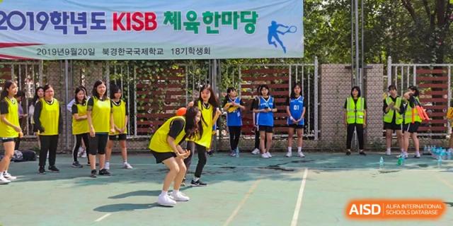 A large group of male students in white shirts and black pants work together during a competitive tug-of-war match on a green sports field at the Korean International School In Beijing.