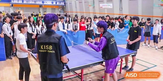Female students in yellow and blue uniforms participate in an outdoor sports match under a large banner featuring the school's logo at the Korean International School In Beijing.
