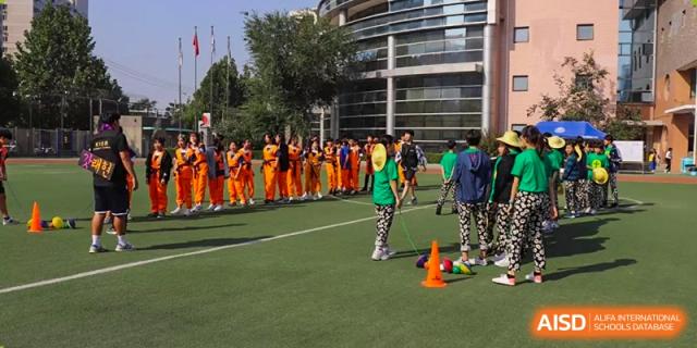 A diverse group of students relaxes and socializes on the sidelines of an outdoor court, with some sitting on the ground at the Korean International School In Beijing.