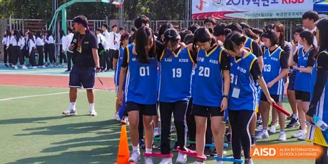 A group of four female students performs an energetic synchronized dance routine in stylish casual wear on an outdoor court at the Korean International School In Beijing.