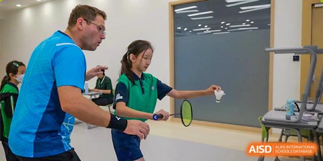 A young student practices her badminton serve while receiving technical guidance from a sports coach during a physical education session at Beijing World Youth Academy.