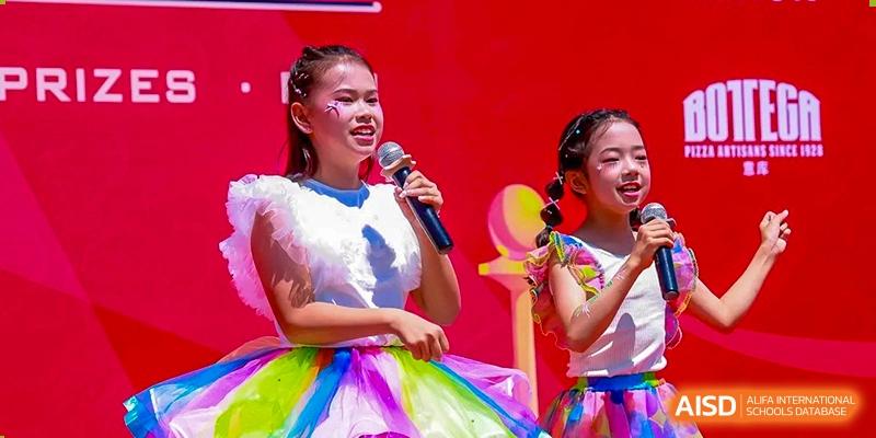 Two young girls in colorful, festive outfits sing into microphones during a stage performance at Beijing World Youth Academy.