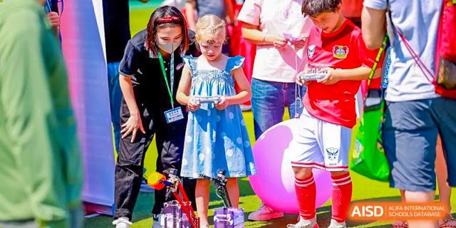 Students and instructors engage in a hands-on technology activity using remote-controlled cars on the grass, fostering teamwork and technical skills outdoors at Beijing World Youth Academy.