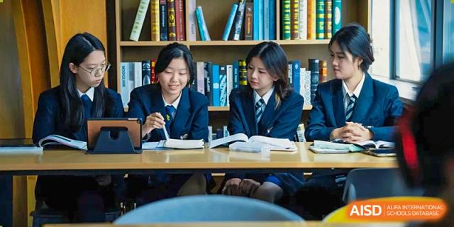 Three students in formal navy blazers sit at a wooden desk and collaborate on a project in front of a full bookshelf at St Bees Dongguan.