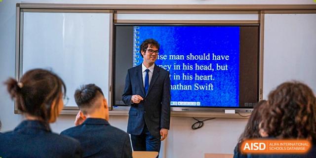 A male teacher in a dark suit stands at the front of a classroom and presents a digital lesson on a large blue screen at St Bees Dongguan.
