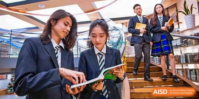 Two female students in formal navy blazers and striped ties review a textbook together on a staircase inside the campus at St Bees Dongguan.