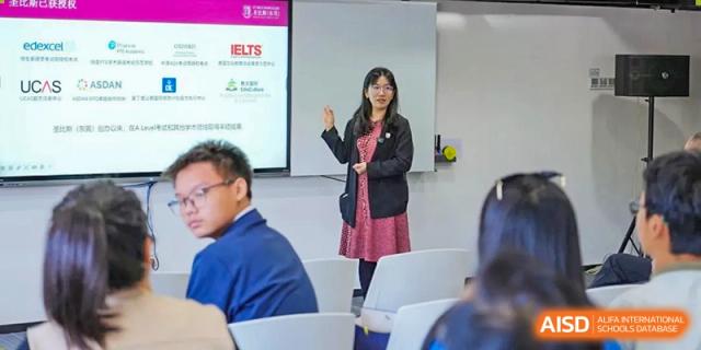 A female teacher stands in front of a digital presentation screen and addresses a group of seated students during a lecture at St Bees Dongguan.