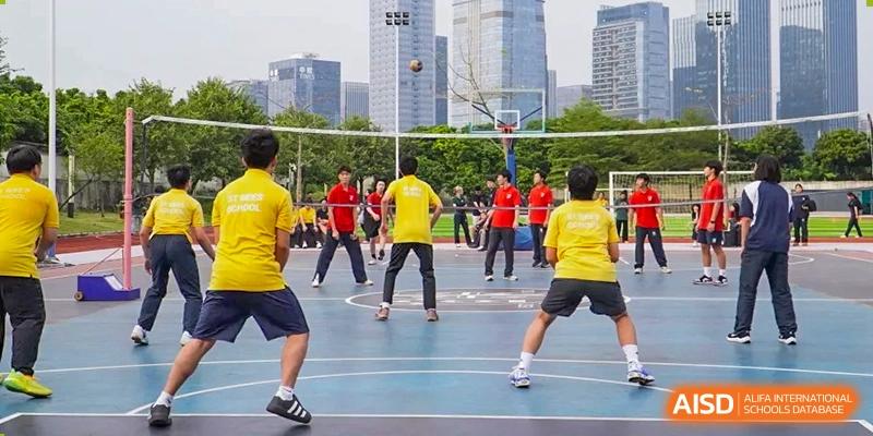 Students in yellow and black jerseys play a competitive game of volleyball on an outdoor blue court surrounded by trees at St Bees Dongguan.