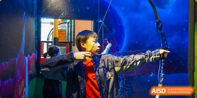 A young student in a red jacket focuses intently while practicing archery with a professional bow against a blue cosmic background at St Bees Dongguan.