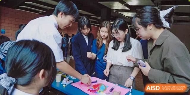 A group of students in formal attire gather around a table to work on a colorful group art or science project at St Bees Dongguan.