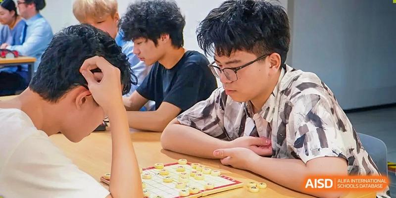A student in a checkered shirt focuses on a board game or puzzle during an indoor recreational session with classmates at St Bees Dongguan.