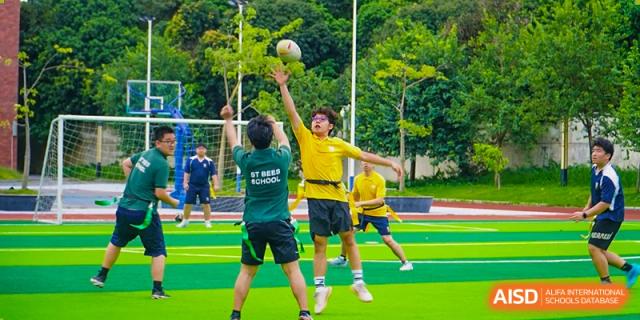 Two students jump high to reach a volleyball during an active outdoor match on a green and blue court at St Bees Dongguan.